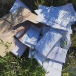 Uma caixa de cartão e vários envelopes e documentos estão espalhados entre a vegetação, ao ar livre. A imagem apresenta o logótipo da TVI.