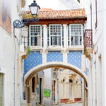 Rua estreita com um edifício que forma um arco sobre a passagem. A fachada tem azulejos azuis e brancos, janelas envidraçadas e um telhado de telha.