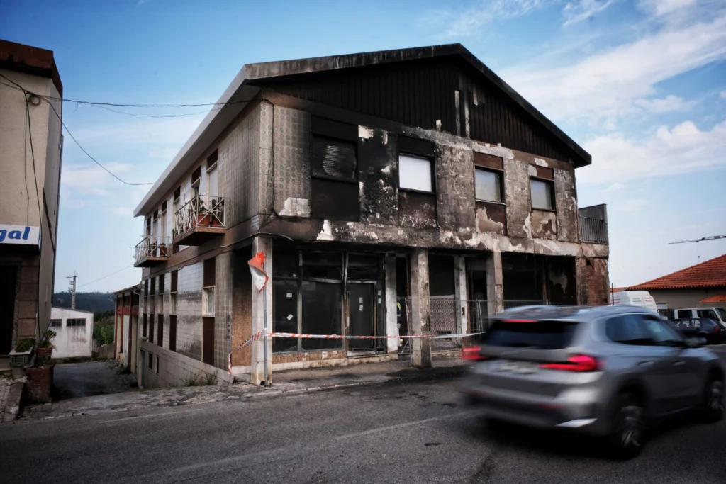 Edifício de dois pisos com a fachada enegrecida e danificada, junto a uma rua. A entrada está delimitada por fita de sinalização e um automóvel passa em frente.