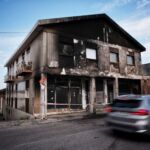 Edifício de dois pisos com a fachada enegrecida e danificada, junto a uma rua. A entrada está delimitada por fita de sinalização e um automóvel passa em frente.