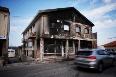 Edifício de dois pisos com a fachada enegrecida e danificada, junto a uma rua. A entrada está delimitada por fita de sinalização e um automóvel passa em frente.