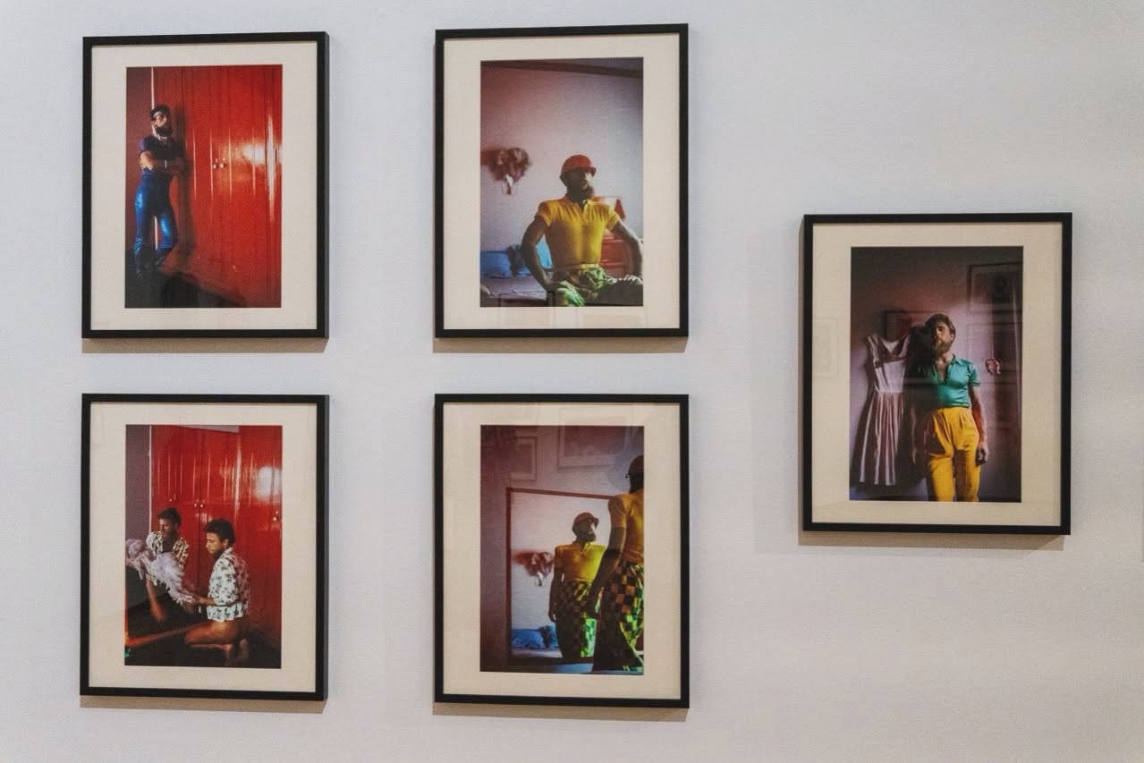 Cinco fotografias emolduradas estão expostas numa parede branca. As imagens mostram pessoas em interiores, com vestuário colorido e portas vermelhas.