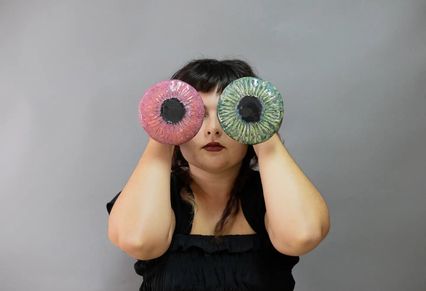 Uma pessoa segura dois objetos circulares coloridos diante dos olhos, um cor-de-rosa e outro verde, com padrões semelhantes a íris. O retrato foi feito perante um fundo cinzento.
