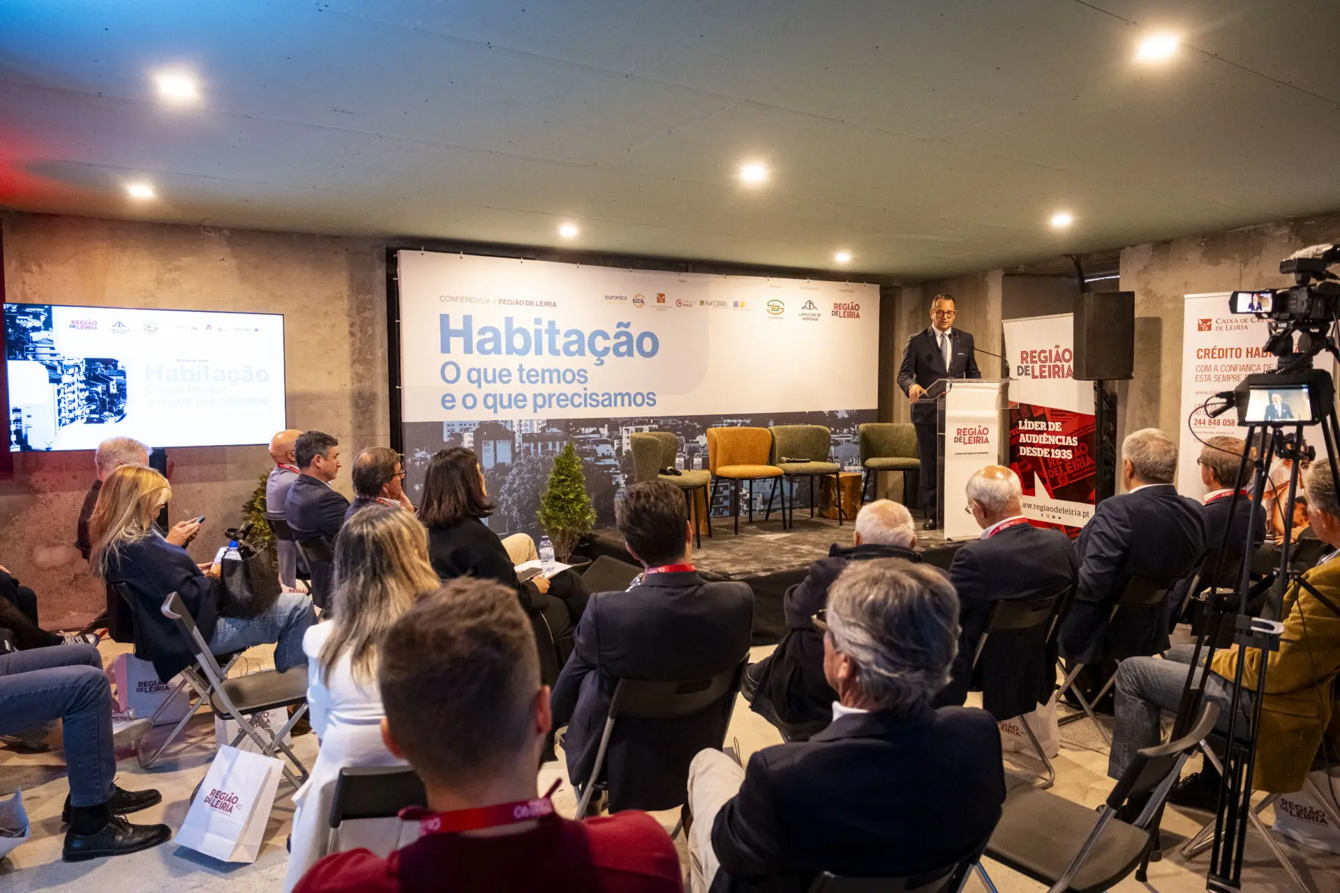 Um orador discursa perante uma plateia numa conferência da Região de Leiria. O painel apresenta o tema «Habitação: O que temos e o que precisamos».