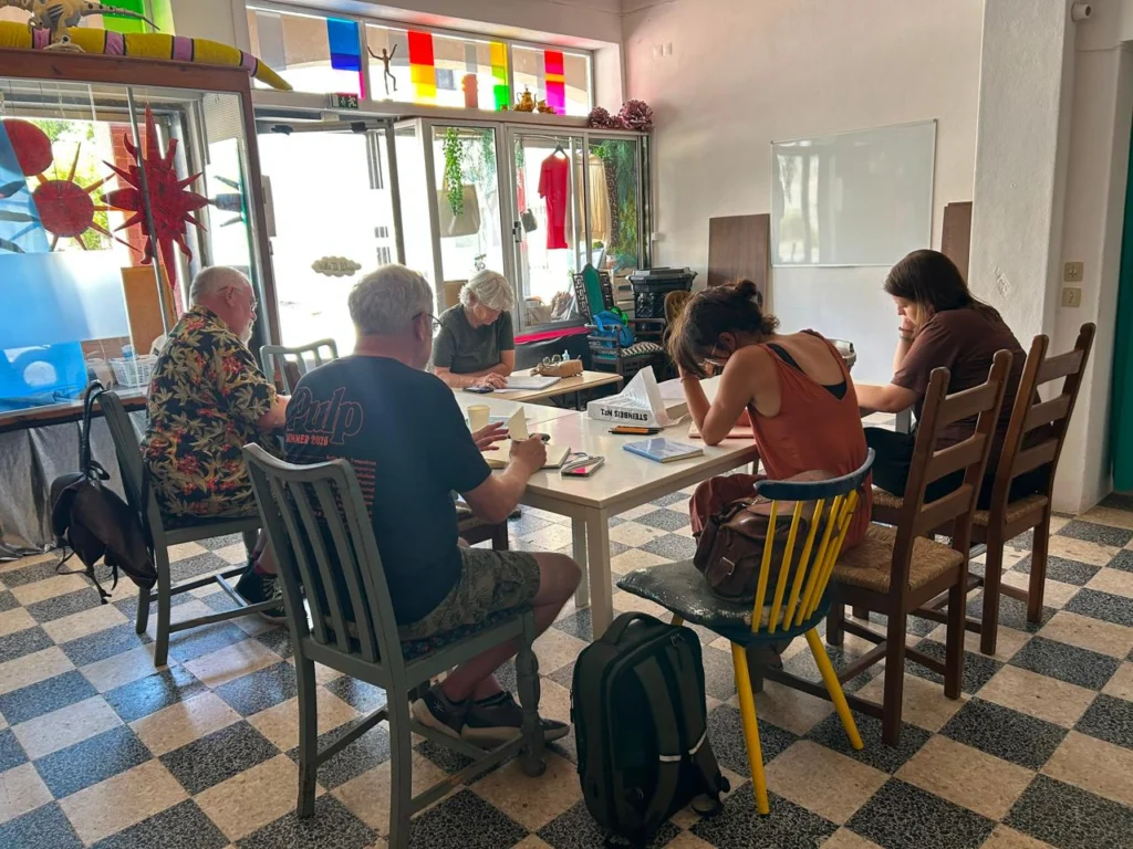 Cinco pessoas estão sentadas à volta de mesas numa sala, aparentemente a escrever ou a consultar apontamentos. O espaço tem janelas com painéis coloridos e um chão em xadrez.