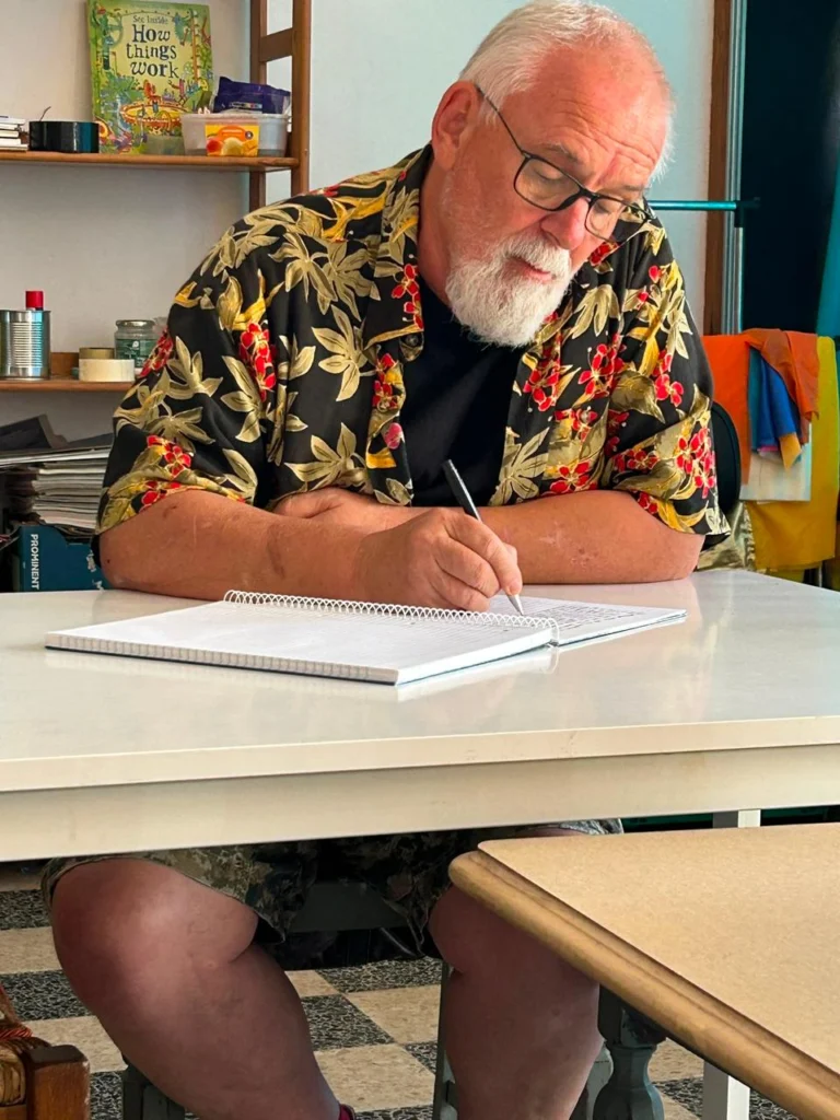 Um homem de cabelo e barba brancos, com óculos, escreve num caderno aberto à mesa. Atrás dele vê-se uma estante com livros e outros objetos.