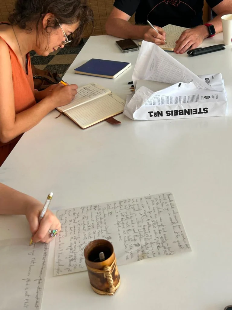 Várias pessoas escrevem à mão em cadernos e folhas, sentadas à volta de uma mesa. Sobre a mesa há documentos, canetas e um telemóvel.