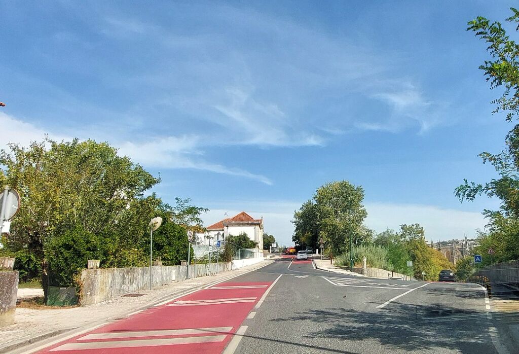 Uma estrada atravessa uma zona arborizada, com passeios, sinalização e veículos ao fundo. Vê-se uma faixa vermelha no pavimento e um edifício de telhado de telha junto à estrada.