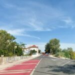 Uma estrada atravessa uma zona arborizada, com passeios, sinalização e veículos ao fundo. Vê-se uma faixa vermelha no pavimento e um edifício de telhado de telha junto à estrada.