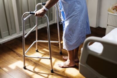 Uma pessoa com bata hospitalar e pulseira no pulso apoia-se num andarilho junto a uma cama. O rosto não está visível.