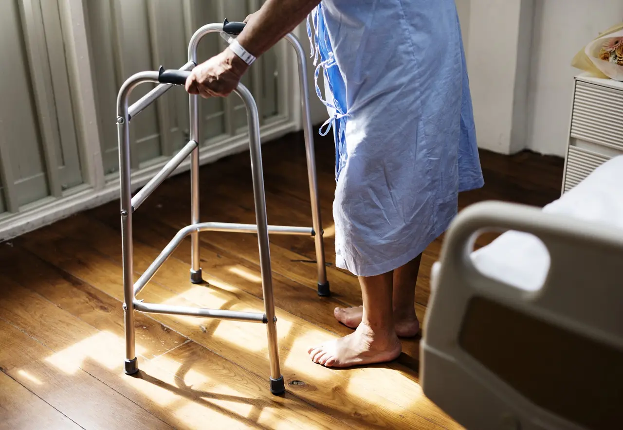 Uma pessoa com bata hospitalar e pulseira no pulso apoia-se num andarilho junto a uma cama. O rosto não está visível.