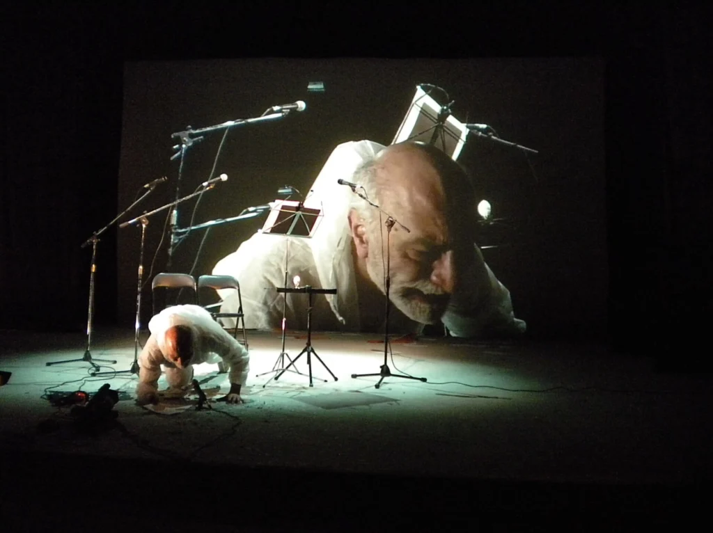 Uma pessoa vestida de branco está apoiada no chão de um palco, diante de vários microfones, cadeiras e estantes de partituras. Ao fundo, uma projeção mostra em grande plano o rosto de um homem.