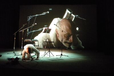 Uma pessoa vestida de branco está apoiada no chão de um palco, diante de vários microfones, cadeiras e estantes de partituras. Ao fundo, uma projeção mostra em grande plano o rosto de um homem.
