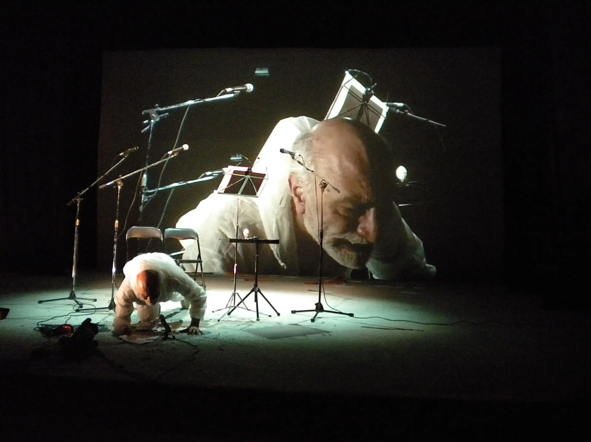 Uma pessoa vestida de branco está apoiada no chão de um palco, diante de vários microfones, cadeiras e estantes de partituras. Ao fundo, uma projeção mostra em grande plano o rosto de um homem.