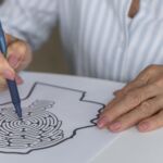 Uma pessoa segura uma caneta sobre uma folha com um labirinto impresso, enquanto apoia a outra mão no papel. O rosto não está visível.