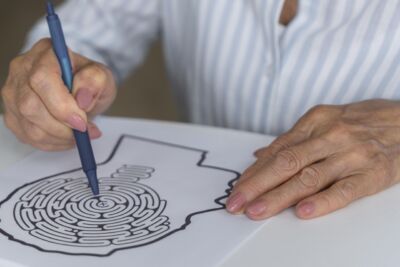 Uma pessoa segura uma caneta sobre uma folha com um labirinto impresso, enquanto apoia a outra mão no papel. O rosto não está visível.