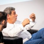 Dois homens estão sentados num espaço interior. O homem em destaque gesticula enquanto fala e segura uma caneta; o outro observa-o.