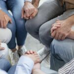 Várias pessoas estão sentadas em círculo e dão as mãos. A imagem mostra apenas partes dos seus corpos; não há rostos nem texto visível.