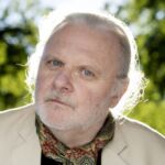 Retrato de um homem de cabelo e barba brancos, com casaco claro e camisola estampada. Está ao ar livre, diante de vegetação desfocada.