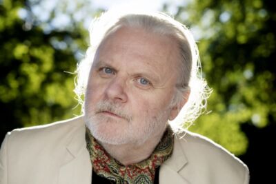 Retrato de um homem de cabelo e barba brancos, com casaco claro e camisola estampada. Está ao ar livre, diante de vegetação desfocada.