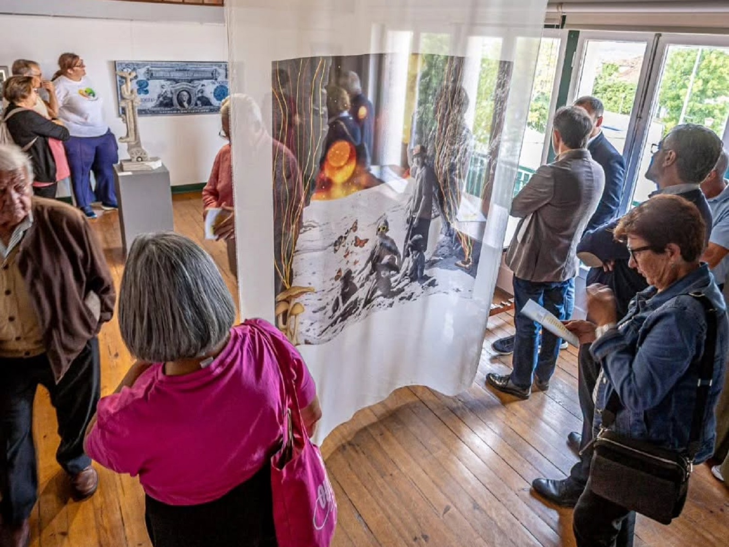 Um grupo de pessoas observa obras numa sala de exposições. Ao centro, uma instalação em tecido translúcido apresenta imagens de uma floresta e de animais.