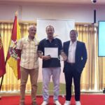 Três homens posam numa sala durante uma cerimónia. O homem ao centro segura um diploma e um troféu; ao fundo, vê-se uma bandeira com a inscrição «Cidade de Albufeira».