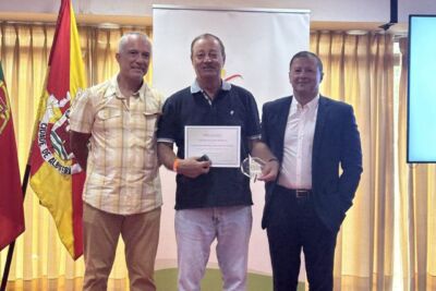 Três homens posam numa sala durante uma cerimónia. O homem ao centro segura um diploma e um troféu; ao fundo, vê-se uma bandeira com a inscrição «Cidade de Albufeira».
