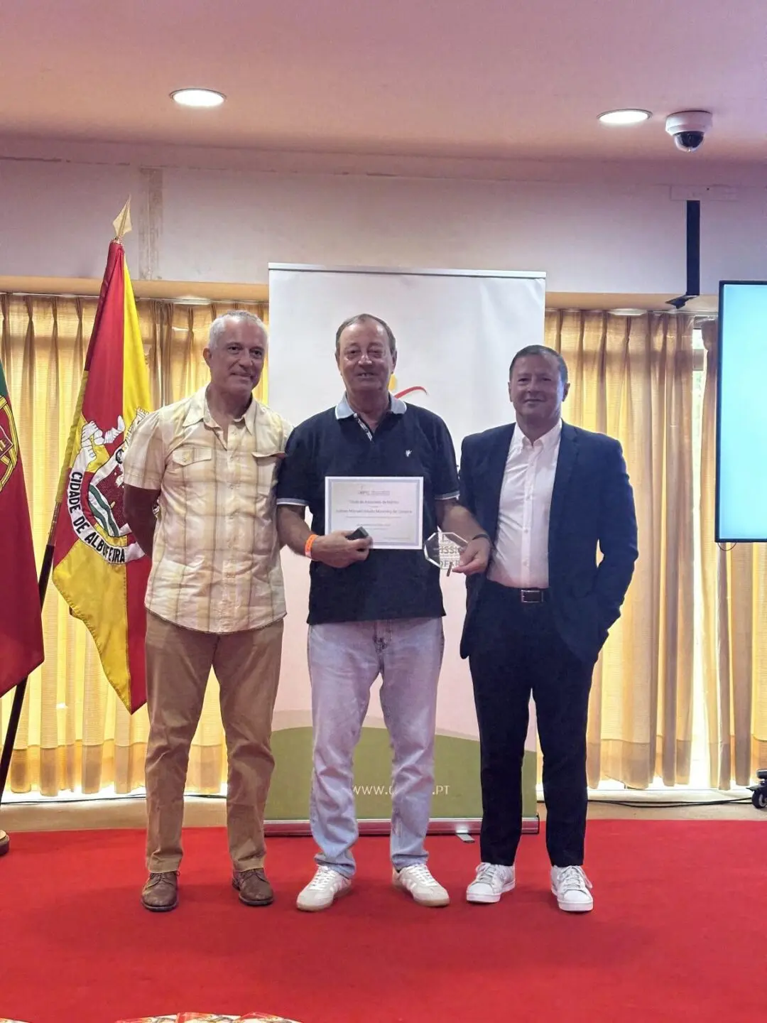 Três homens posam numa sala durante uma cerimónia. O homem ao centro segura um diploma e um troféu; ao fundo, vê-se uma bandeira com a inscrição «Cidade de Albufeira».