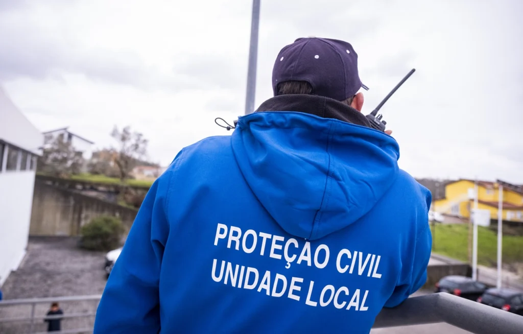 Uma pessoa vista de costas, com um casaco azul da Proteção Civil — Unidade Local, utiliza um rádio portátil no exterior.
