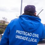 Uma pessoa vista de costas, com um casaco azul da Proteção Civil — Unidade Local, utiliza um rádio portátil no exterior.