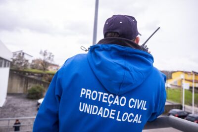 Uma pessoa vista de costas, com um casaco azul da Proteção Civil — Unidade Local, utiliza um rádio portátil no exterior.