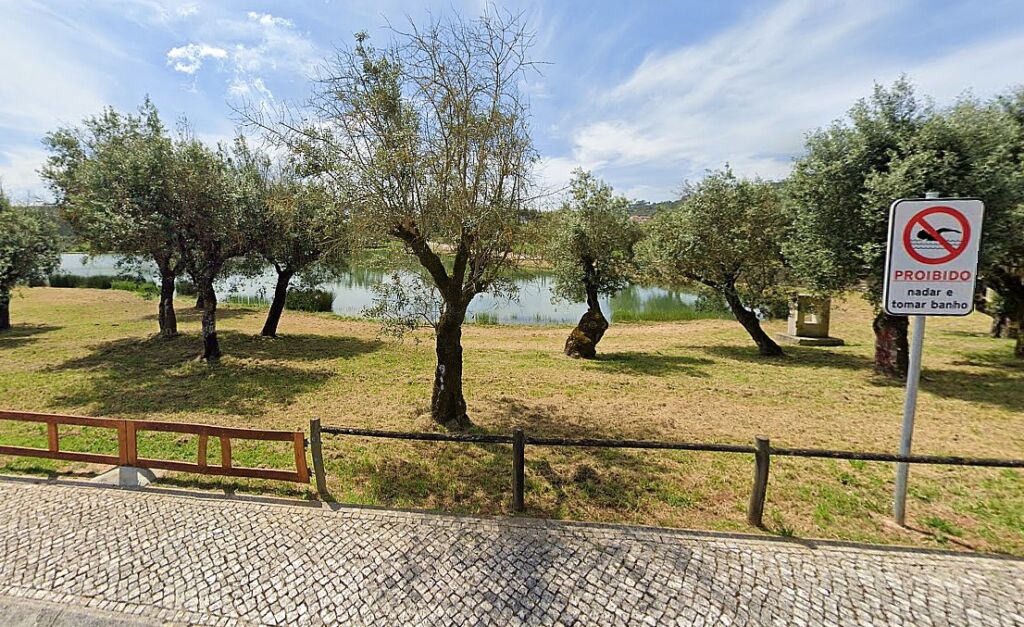 Zona arborizada junto a um plano de água, vista de um passeio em calçada. Um sinal indica que é proibido nadar e tomar banho.