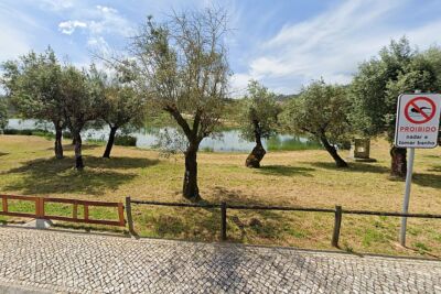 Zona arborizada junto a um plano de água, vista de um passeio em calçada. Um sinal indica que é proibido nadar e tomar banho.