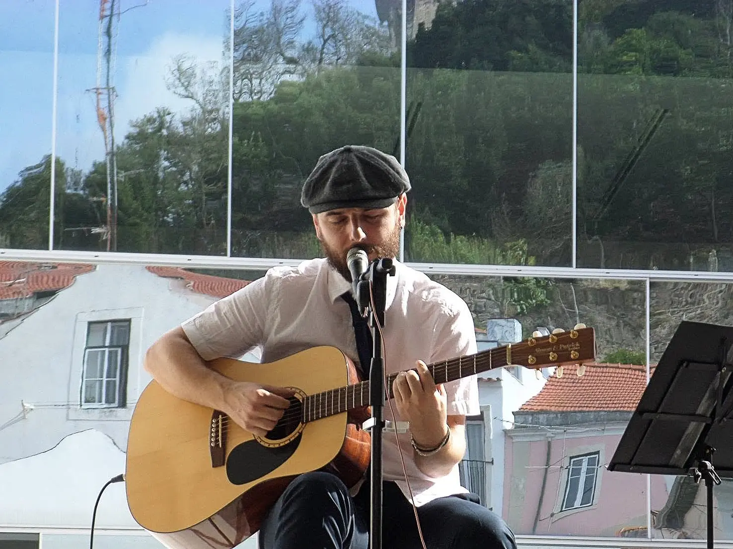 Um músico canta ao microfone enquanto toca guitarra acústica. A atuação decorre num espaço envidraçado, com edifícios e vegetação visíveis ao fundo.