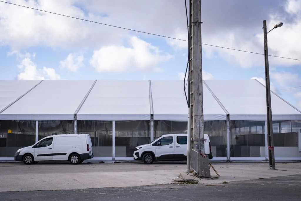 Uma grande estrutura coberta por uma lona branca ocupa o fundo da imagem. Em frente, estão estacionadas duas carrinhas brancas, junto a postes e cabos elétricos.