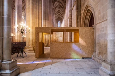Interior de uma igreja de arquitetura gótica, com colunas de pedra, abóbadas, vitrais e bancos. Uma estrutura temporária de painéis de madeira ocupa parte da nave.