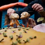 Um manipulador movimenta duas marionetas de tecido num cenário colorido com pequenas plantas e um grande objeto cor-de-rosa. A imagem mostra um momento de teatro de marionetas.