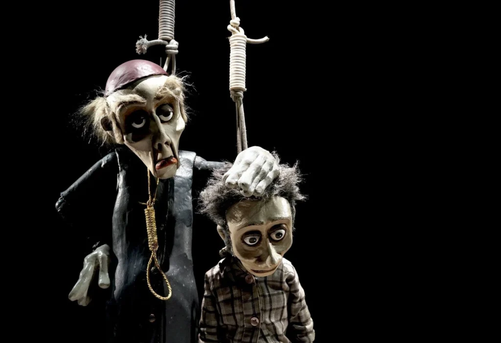 Dois bonecos de marionetas estão suspensos por cordas sobre um fundo preto. O boneco maior, vestido de preto, pousa a mão na cabeça do mais pequeno, que veste uma camisola aos quadrados.