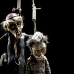 Dois bonecos de marionetas estão suspensos por cordas sobre um fundo preto. O boneco maior, vestido de preto, pousa a mão na cabeça do mais pequeno, que veste uma camisola aos quadrados.