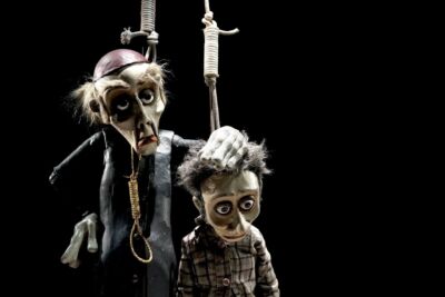 Dois bonecos de marionetas estão suspensos por cordas sobre um fundo preto. O boneco maior, vestido de preto, pousa a mão na cabeça do mais pequeno, que veste uma camisola aos quadrados.