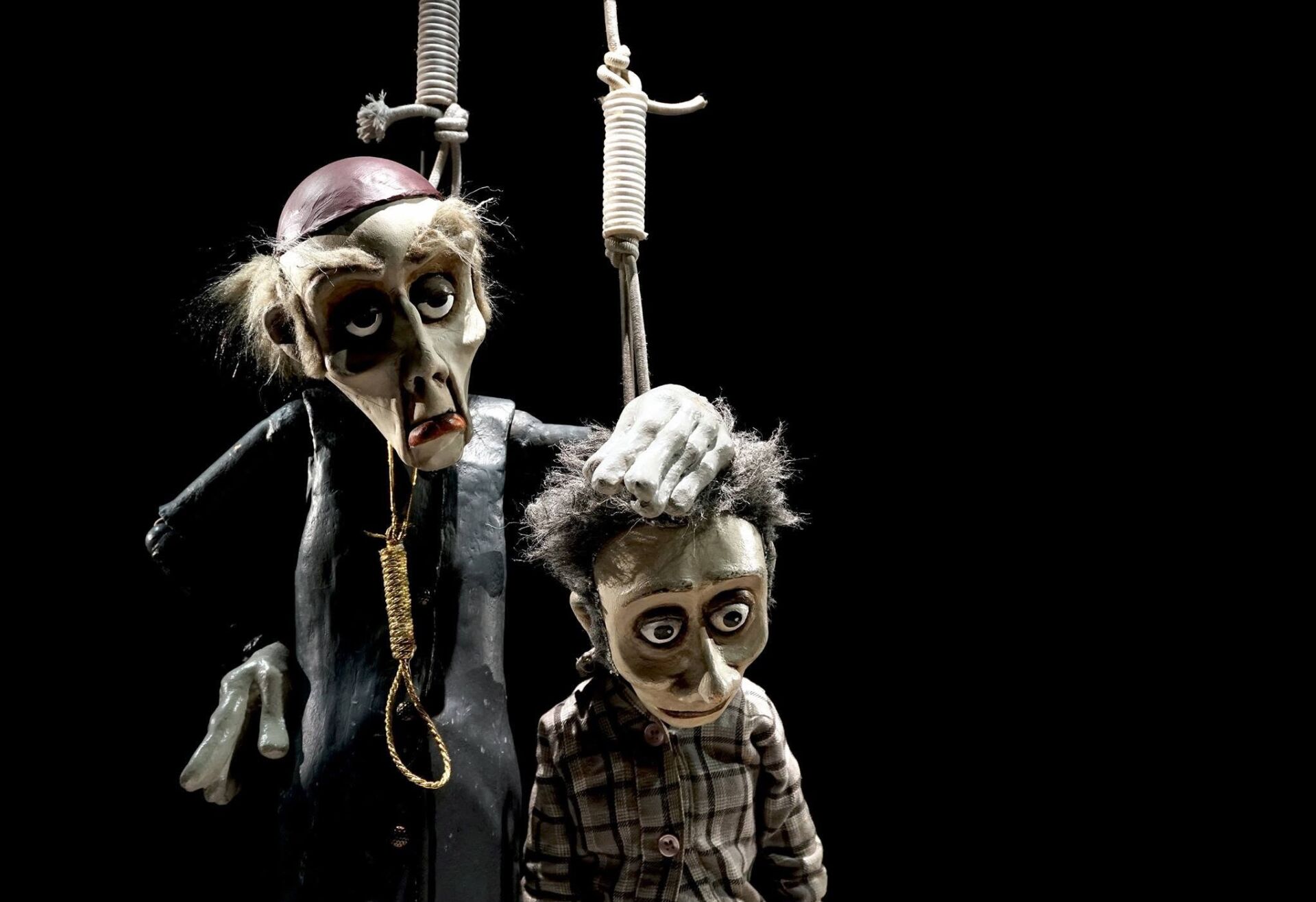 Dois bonecos de marionetas estão suspensos por cordas sobre um fundo preto. O boneco maior, vestido de preto, pousa a mão na cabeça do mais pequeno, que veste uma camisola aos quadrados.