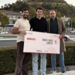 Três pessoas posam com um cartaz do Hackathon Climate U que apresenta a mensagem «Duas idas COP31». Ao fundo, vê-se um castelo numa colina.