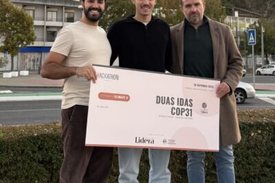 Três pessoas posam com um cartaz do Hackathon Climate U que apresenta a mensagem «Duas idas COP31». Ao fundo, vê-se um castelo numa colina.