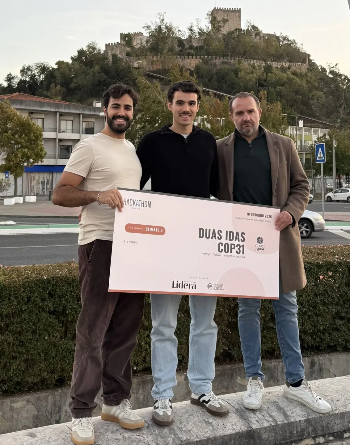 Três pessoas posam com um cartaz do Hackathon Climate U que apresenta a mensagem «Duas idas COP31». Ao fundo, vê-se um castelo numa colina.