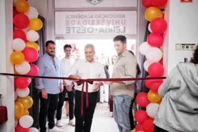Um homem prepara-se para cortar uma fita numa cerimónia à entrada de um espaço decorado com balões vermelhos, brancos e dourados. Na porta lê-se, de forma invertida, «Associação Académica Universidade Leiria • Oeste».