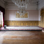 Sala ampla e vazia, com chão de madeira, estrado ao fundo e lustres de cristal. As paredes têm painéis decorativos, cortinas vermelhas e fotografias emolduradas.