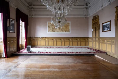 Sala ampla e vazia, com chão de madeira, estrado ao fundo e lustres de cristal. As paredes têm painéis decorativos, cortinas vermelhas e fotografias emolduradas.