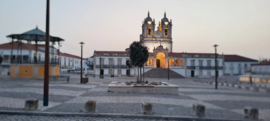 Uma igreja com duas torres sineiras e um relógio está iluminada ao entardecer. Em frente, vê-se uma praça empedrada praticamente vazia, com árvores e bancos.