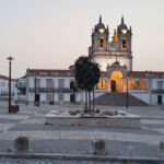 Uma igreja com duas torres sineiras e um relógio está iluminada ao entardecer. Em frente, vê-se uma praça empedrada praticamente vazia, com árvores e bancos.