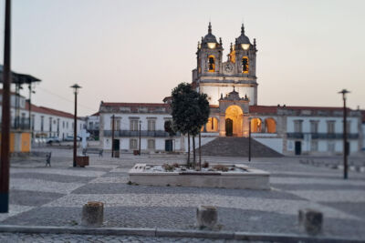 Uma igreja com duas torres sineiras e um relógio está iluminada ao entardecer. Em frente, vê-se uma praça empedrada praticamente vazia, com árvores e bancos.